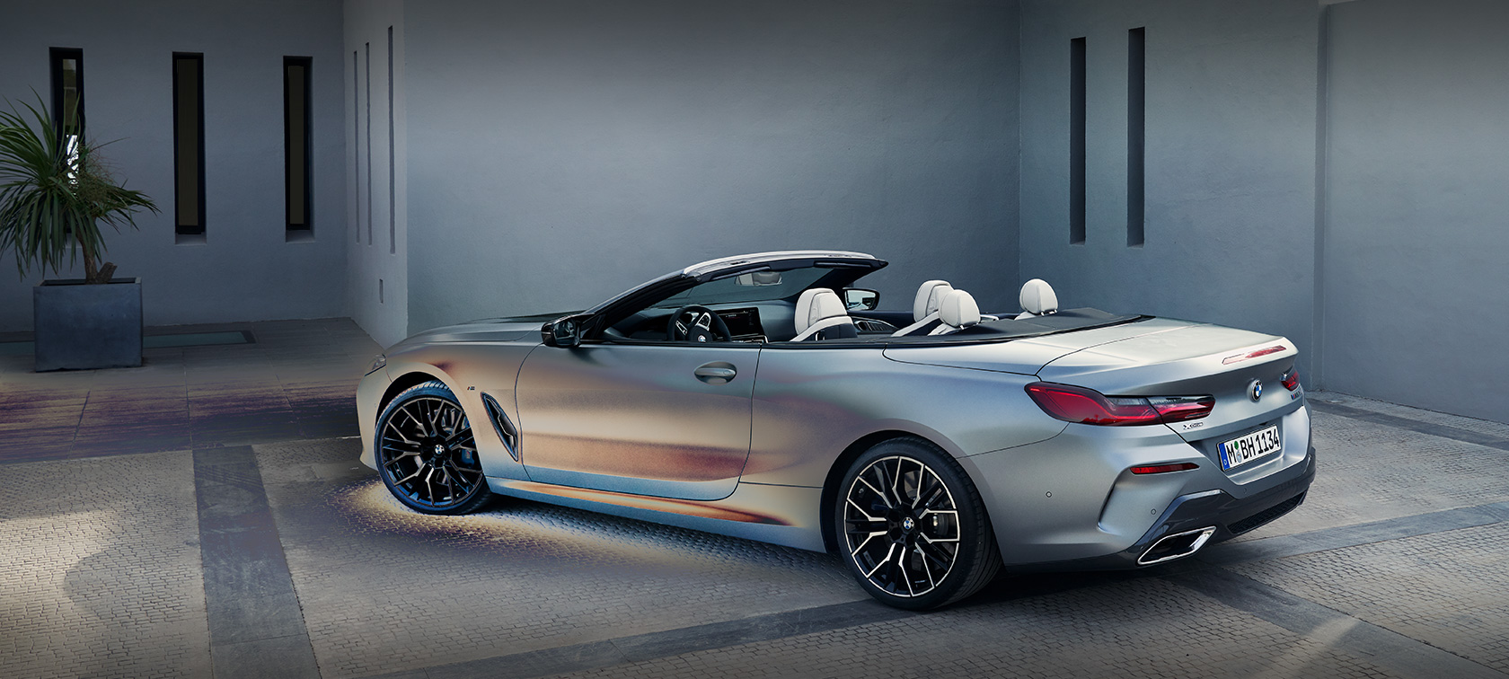 Мужчина прислонился к неподвижному BMW 8 серии Cabrio G14 LCI (рестайлинг 2022 года) в эксклюзивном цвете BMW Individual Чистый Морозный Серый металлик, стоящему на фоне здания, вид сбоку