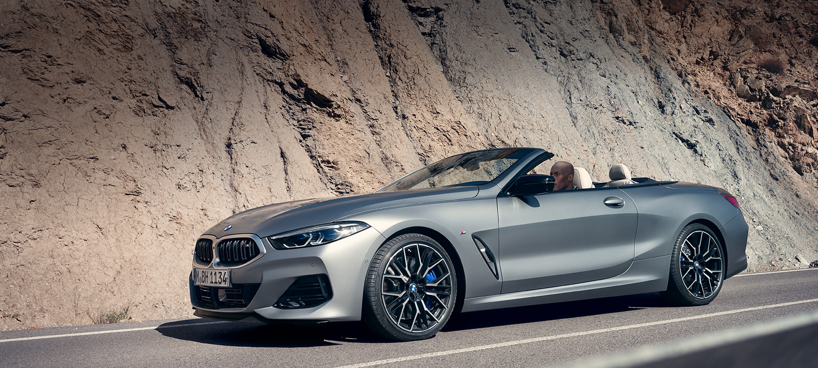 BMW 8 серии Cabrio G14 LCI (рестайлинг 2022 года) с убранным мягким верхом в движении на фоне отвесной скалы BMW 8 серии Cabrio G14 LCI (рестайлинг 2022 года) эксклюзивного цвета BMW Individual Морозный Серый металлик с убранным мягким верхом в движении на фоне отвесной скалы, вид сбоку