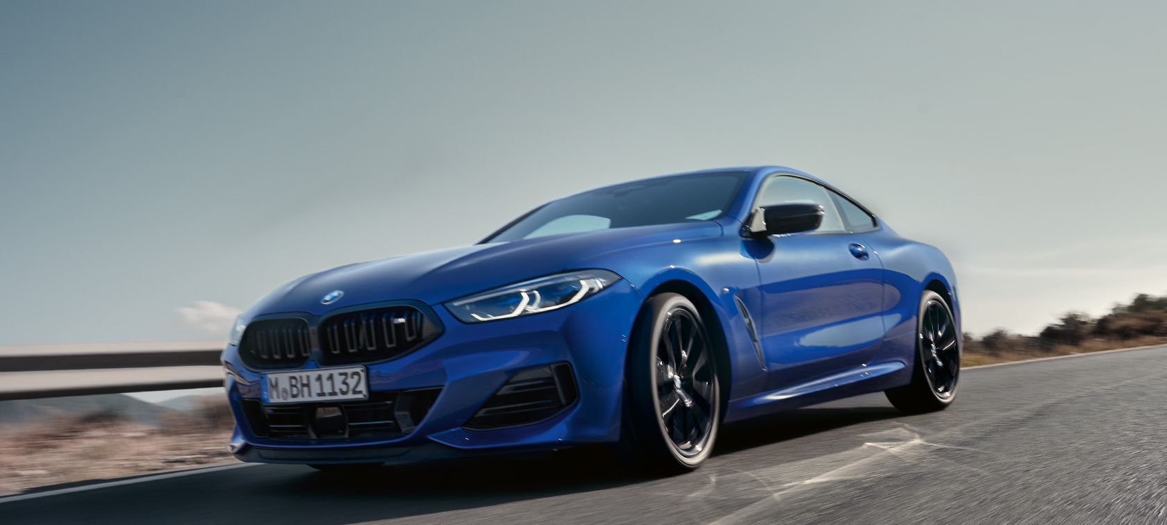 BMW 8 серии Coupé G15 LCI (рестайлинг 2022 года) BMW M850i xDrive M цвета Синий Портимау металлик вид спереди, съемка с низкой точки BMW 8 серии Coupé G15 LCI (рестайлинг 2022 года) BMW M850i xDrive M цвета Синий Портимау металлик движется по загородной дороге на фоне холмов, вид спереди, съемка с низкой точки
