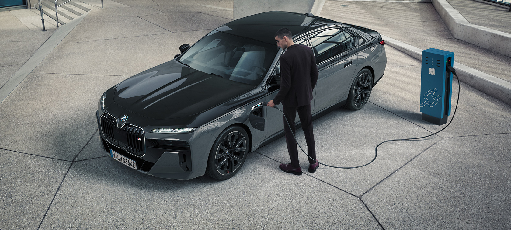 BMW M760e xDrive G70, пакет M Performance BMW M760e xDrive G70, пакет M Performance, BMW Individual двухцветная окраска "Черный Сапфир металлик" и "BMW Individual Серый Дравит металлик", вид спереди в три четверти, мужчина с зарядным кабелем рядом с зарядной станцией