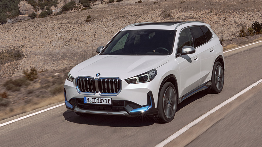 BMW iX1 U11 fпервого поколения, вид спереди с поворотом в три четверти, в движении по дороге BMW X Серии BMW iX1 U11 первого поколения, вид спереди с поворотом в три четверти, в движении по дороге